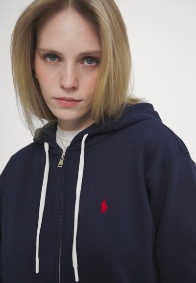 Sweatshirt azul marinho com capuz, cordões brancos, fecho de correr frontal e um logótipo vermelho no peito. Textura suave, design casual.