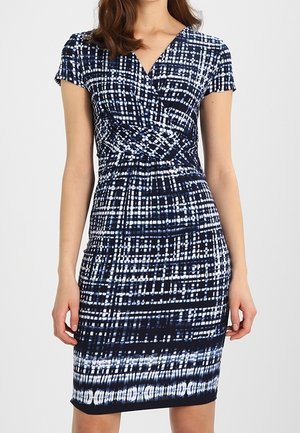 Kobieta ubrana w sukienkę do kolan z krótkim rękawem, w czarno-biały abstrakcyjny wzór w kratę, z dekoltem w serek.
