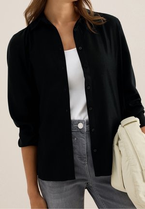 Femme portant une chemise noire boutonnée sur un haut blanc avec un jean gris, tenant une veste beige pliée devant un fond uni.