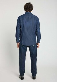 Chemise en denim à boutons avec des manches longues, couleur bleu foncé, détail de bas de dos et coupe ajustée. Portée avec un pantalon assorti de couleur sombre et des chaussures noires.