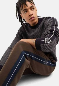 Maglione in bianco e nero testurizzato con motivo a maglia, dotato di cuciture a contrasto e patch con logo. Pantaloni marroni con strisce e dettagli in blu navy.