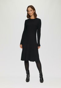Zwarte gebreide jurk met lange mouwen, ronde hals en een uitlopende zoom. Gemaakt met zwarte maillots en enkellaarsjes. Soepele textuur.