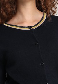 Navy Cardigan mit Rundhalsausschnitt und geripptem Streifenrand in Gelb, Blau und Lavendel. Fünf Knopf-Frontverschluss. Weicher Stoff.