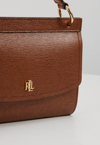 Hnědá kůže texturovaná kabelka s klopovým uzavřením. Obsahuje zlatý logo detail a popruh s kovovými prvky.