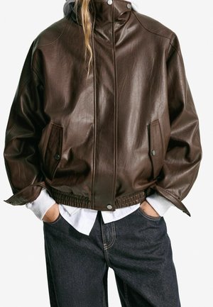 Person trägt dunkelbraune Lederjacke mit hohem Kragen, weißes Hemd darunter und dunkle Jeans, Hände in den Taschen vor einfachem Hintergrund.
