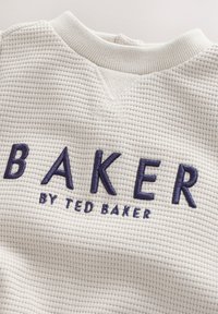 Vitt, texturerat tyg med ett ribbat mönster som har "BAKER" i marinblå broderi och "by TED BAKER" i mindre text nedanför.