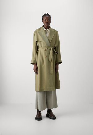 Manteau trench double boutonnage vert olive avec une ceinture, doté d'un large col et de manches longues. Porté par-dessus un pantalon large gris clair et des bottes marron.