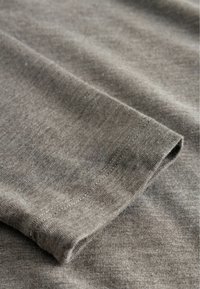 Pelēka kokvilnas auduma ar gludu tekstūru, kuram ir salocīta mala un kārtīgas šuves. Redzamas smalkas krāsas un raksta variācijas.