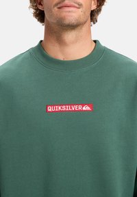 Muž v zelené mikině s červenou obdélníkovou nášivkou, na které je logo a text "QUIKSILVER", na hrudi, na bílém pozadí.
