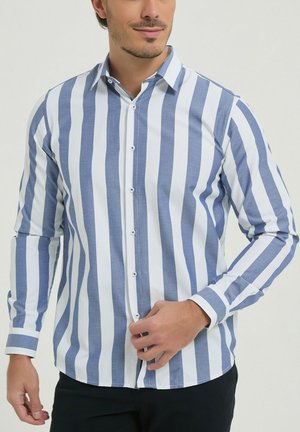 Homme portant une chemise à manches longues à rayures verticales bleues et blanches, boutonnant le bouton du bas, sur un fond uni.