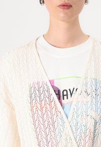 Cardigan crem din dantelă, cu un model geometric, purtat peste un tricou alb cu grafică, având accente colorate și text.
