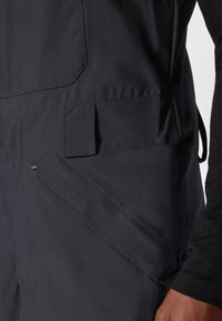 Gros plan d'une personne portant un pantalon noir avec de grandes poches latérales inclinées et une manche longue noire visible.