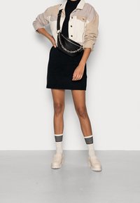 Giacca di jeans color block in crema e grigio sopra un vestito a maglia nero. Borsa a tracolla nera con catena. Scarpe beige e calzini a righe.