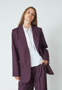 Temno vijoličast blazer z ostrimi reverji, enim sprednjim žepom in ohlapnim krojem, v kombinaciji z belim srajčnim zgornjim delom.