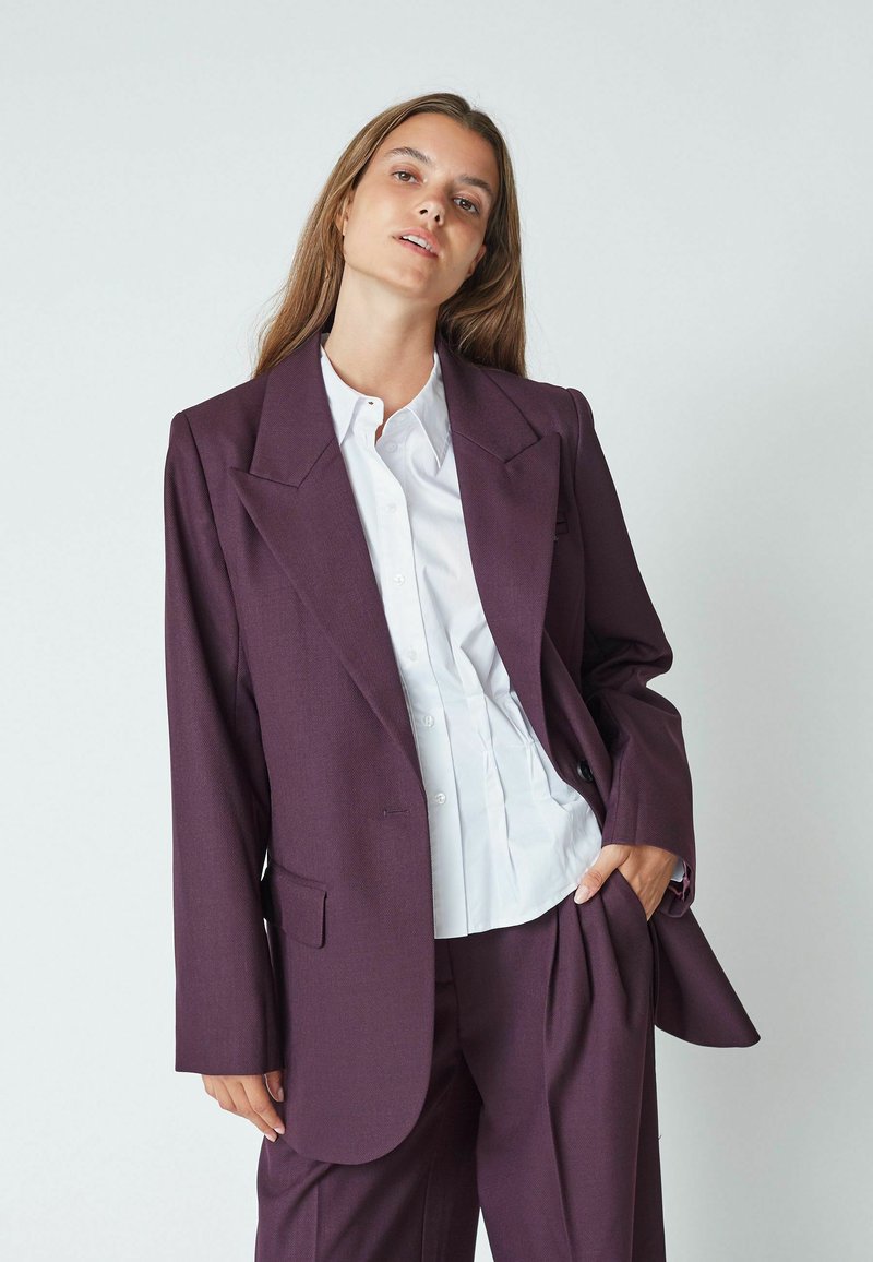 Blazer viola scuro con revers affilati, tasca frontale singola e vestibilità ampia, abbinato a una camicia bianca con bottoni sotto.