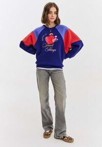 Niebieska, czerwona i fioletowa bluzka z kolorowymi blokami, z graficznym napisem i motywem owoców. Połączona z szarymi dżinsami w stylu dzwonów i butami w zwierzęcy wzór.