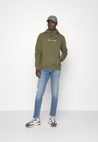 Hoodie oliivinvihreänä, jossa valkoinen logo, kengurupussitasku. Yhdistetty vaaleansinisiin farkkuihin ja harmaaseen lippikseen. Kengissä on tummansinisiä ja kermavalkoisia yksityiskohtia.