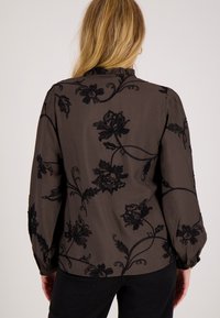 Vrouw met lang blond haar, gekleed in een donkerblouse met bloemenpatroon en lange mouwen, en zwarte broek, met haar rug naar de camera toe.