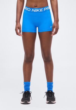 Atleta vistiendo pantalones cortos Nike Pro azul brillante, calcetines azules a juego y zapatillas de correr negras, de pie contra un fondo blanco.