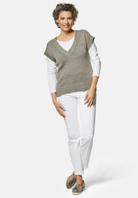 Lichte, grijze gebreide trui met een diepe V-hals en korte mouwen, gedragen over een wit longsleeve shirt, gecombineerd met witte broek en met een patroon versierde schoenen.