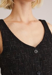 Črna obleka z teksturo, ki ima V-izrez, dve gumb na sprednji strani in vzorec subtilnih sijočih niti, kar ustvarja vizualno globino.