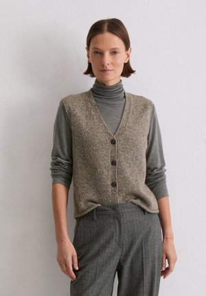 Braune, strukturierte Strickweste mit V-Ausschnitt und dunklen Knöpfen, getragen über einem eng anliegenden grauen Rollkragenpullover. Graue karierte Hosen vervollständigen das Outfit.