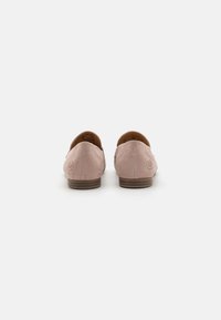 Des chaussures slip-on en suède rose avec une texture lisse, un dos arrondi et un petit talon marron, mettant en avant un logo discret en relief sur chaque côté extérieur.
