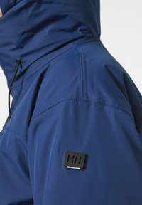 Chaqueta impermeable de color azul marino con un alto cuello, capucha ajustable con cordón y un parche de goma con el logo en el hombro izquierdo.