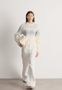 Mulher a caminhar vestindo um suéter creme com padrão, calças brancas de perna larga e segurando uma carteira creme dobrada contra um fundo branco liso.