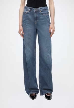 Personne portant un jean bleu en denim ample et des chaussures noires à bout pointu, debout devant un fond clair uni.