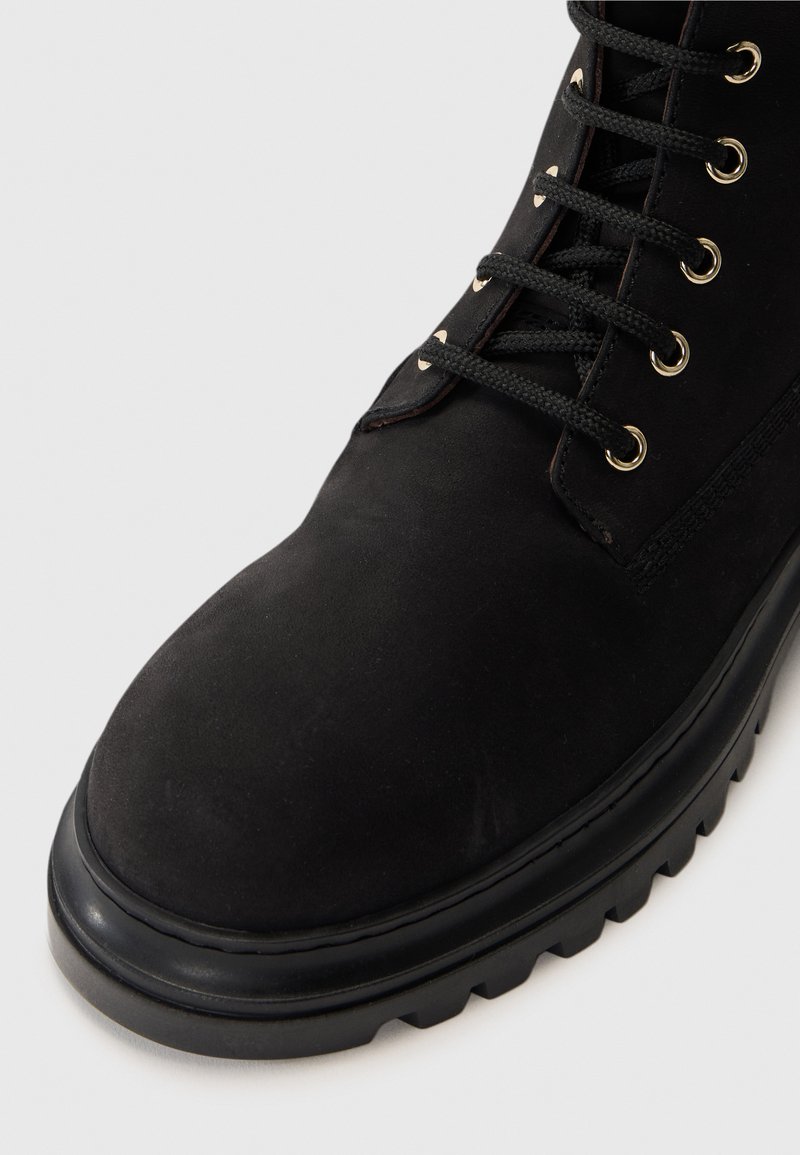 Svart mocka ankelboot med rund tå, texturerad yta, gyllene öglor och svarta snören; har en kraftig svart sula.