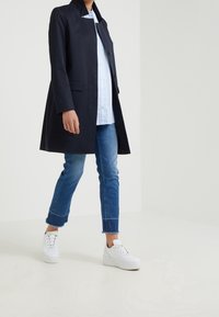 Abrigo azul marino con cuello y un solo botón, combinado con una camisa de botones de color azul claro y jeans azules desgastados. Zapatillas blancas completan el conjunto.