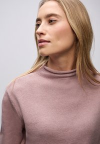 Vaaleanpunainen neulepusero, jossa on korkea, käännettävä kaula. Silkkinen pinta ja rento malli. Hiukset valahtavat pehmeästi olkapäälle. Neutraali tausta.