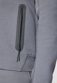 Felpa grigia con un design aderente, dotata di una tasca con zip sul maniche. Texture liscia e cuciture pulite sono ben visibili.
