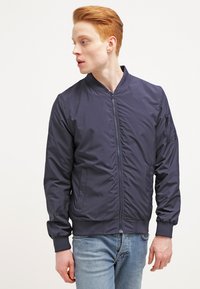 Jachetă de tip bomber navy cu fermoar frontal, manșete și margine cu nervuri. Dispune de un buzunar lateral cu fermoar; fabricată dintr-un material neted, ușor.