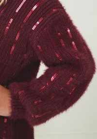 Vijoličen pulover z mehko teksturo, okrašen z navpičnimi vrstami sijočih rdečih sequins na dolgih rokavih.