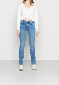 Žena v světle modrých skinny džínech, bílém topu s dlouhým rukávem a volánky, a bílých kotníkových botách s kovovými špičkami, stojící na jednoduchém pozadí.