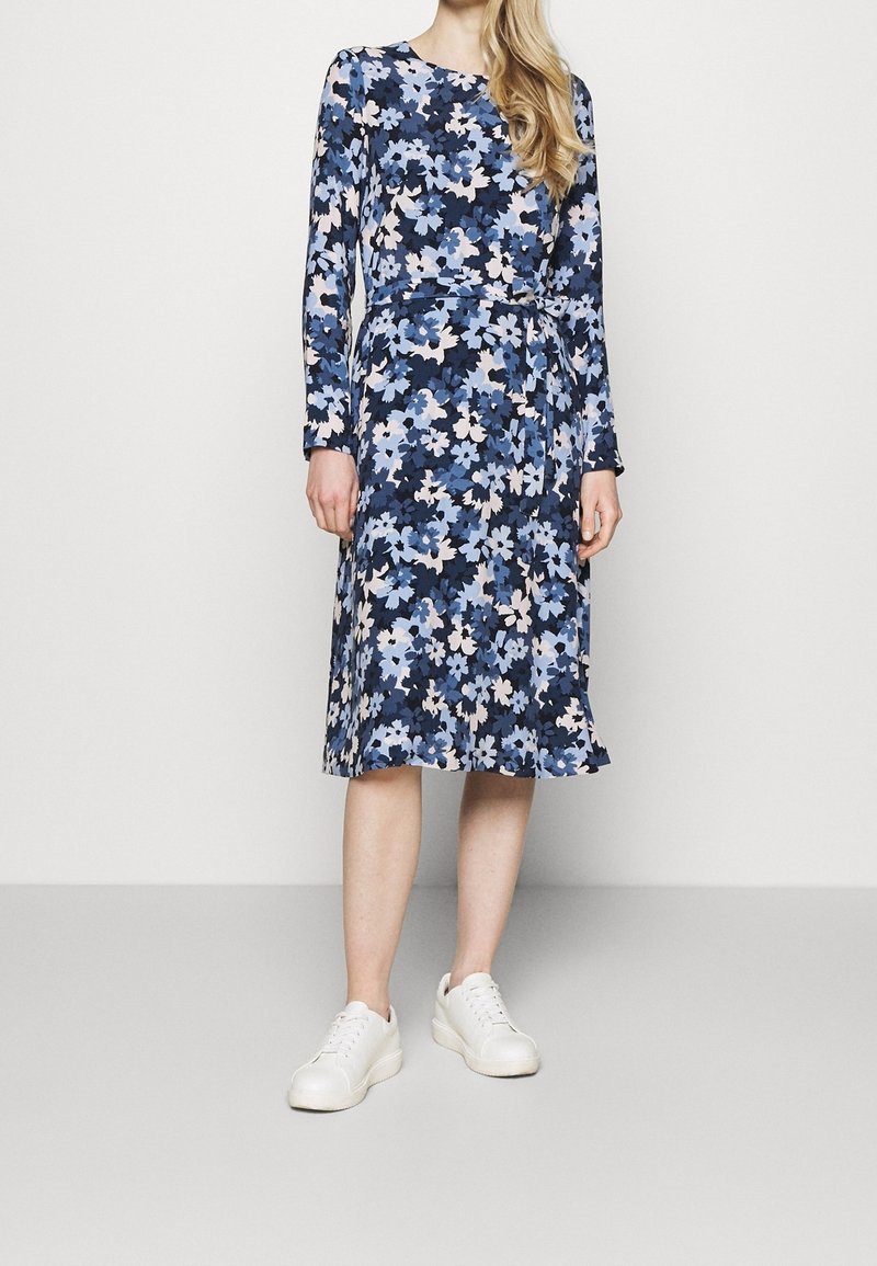 Femme portant une robe bleue à fleurs longueur genou avec manches longues et ceinture à la taille, associée à des baskets blanches, debout devant un fond uni.