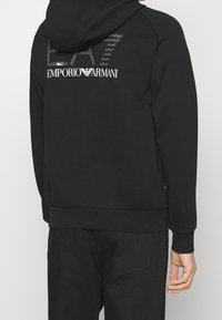 Svart huvtröja med en strukturerad logotyp i vitt på baksidan, ribbade ärmslut och nederkant, med en slät yta.