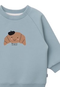 Ljusblå sweatshirt med en croissant-tecknad figur, prydd med en svart basker, som har ett glatt ansikte och texten "Bonjour mon amour."