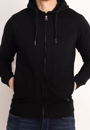 Mężczyzna w czarnej bluzie na zamek z kapturkiem i sznurkami, z rękami w kieszeniach, ubrany w niebieskie dżinsy na jasnym, jednolitym tle.