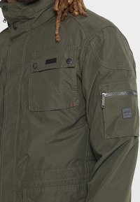 Veste vert olive avec un col haut, plusieurs poches avant, une poche zippée sur la manche et un tissu texturé pour plus de durabilité.