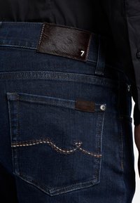 Mörkblå denimjeans med en strukturerad svart läderpatch i midjan med siffran 7, som framhäver kontrastsömmar på fickorna.