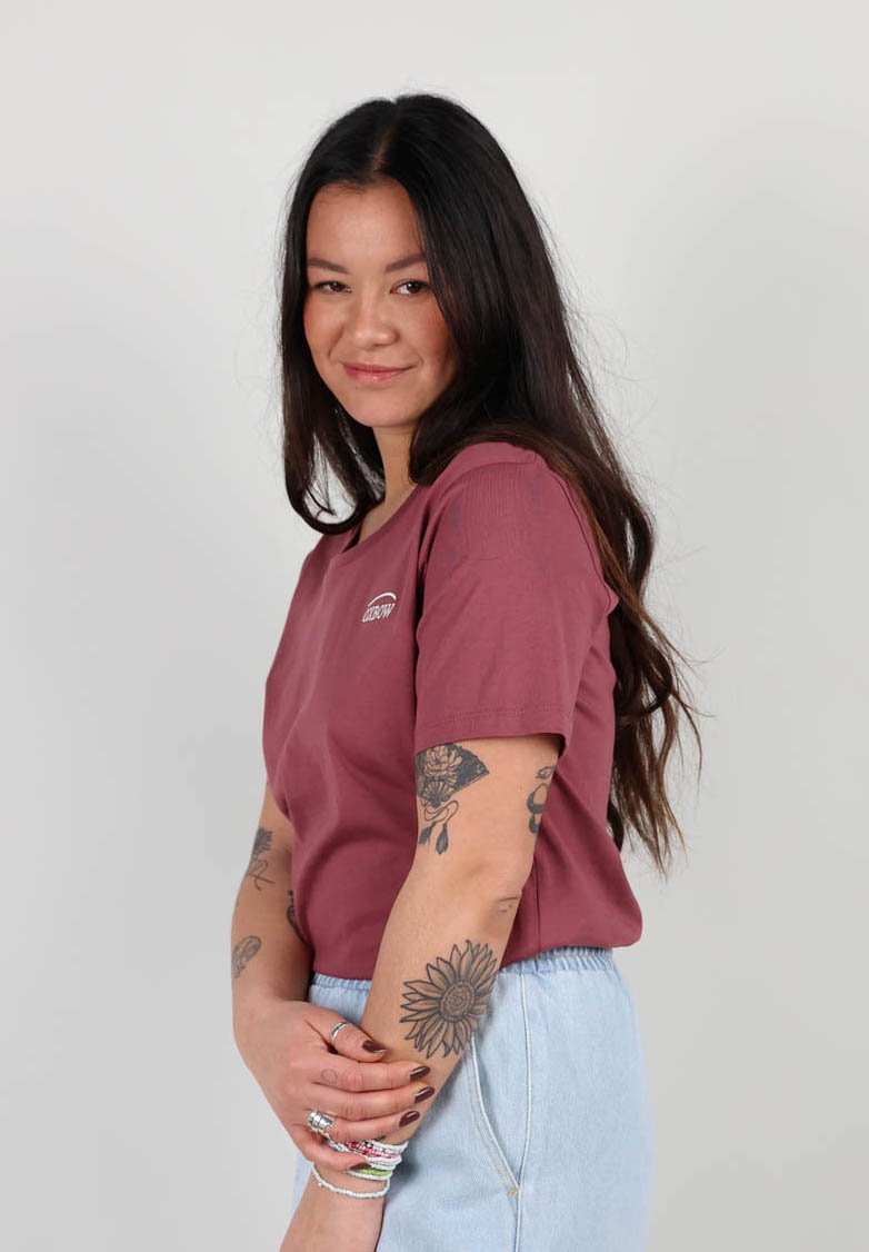 T-shirt bordeaux à manches courtes avec un col rond et un logo brodé, associé à un short en denim bleu clair ; le modèle a des tatouages et des bracelets.