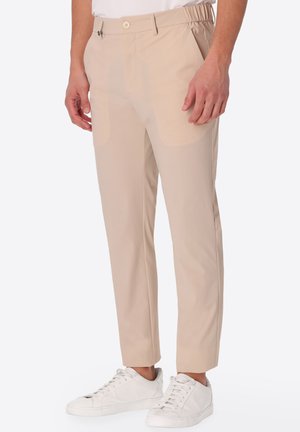 Uomo che indossa pantaloni beige slim fit con chiusura a bottoni e sneakers bianche su sfondo bianco.