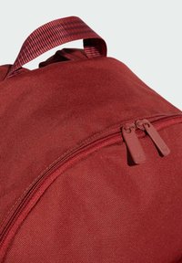 Zaino in tessuto rosso con una superficie liscia, dotato di chiusura con doppia cerniera e manico superiore. Design minimalista senza loghi visibili.