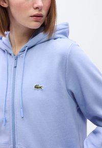 Φωτεινό γαλάζιο φούτερ με φερμουάρ από απαλό ύφασμα, με πράσινο λογότυπο κροκόδειλου στο στήθος και κουκούλα με κορδόνι για ρύθμιση.