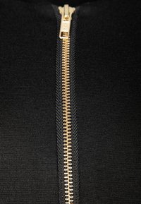 Svart tyg närbild med en guldfärgad dragkedja, med synliga tänder och söm detaljer längs dragkedjan. Slät textur.