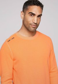 Een oranje sweatshirt met ronde hals, gemaakt van gestructureerde stof, met een schouderaccent met de tekst "CAMP COVID." Eenvoudig, hedendaags ontwerp.