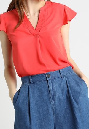 Mujer con blusa coral de manga corta metida dentro de pantalones vaqueros azul de talle alto con pliegues, con las manos en los bolsillos.